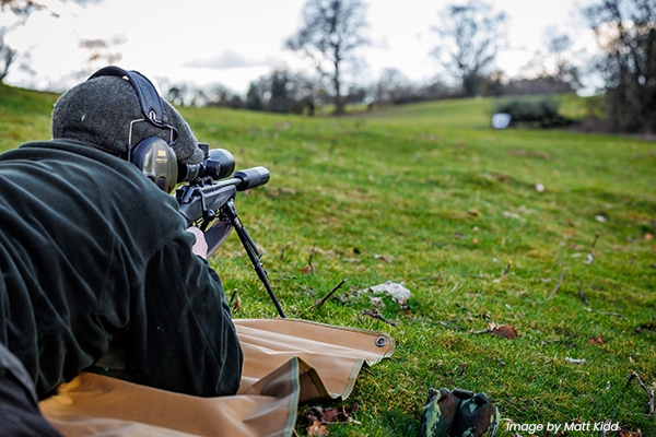 A deer stalking lying down aiming through the scope of their rifle